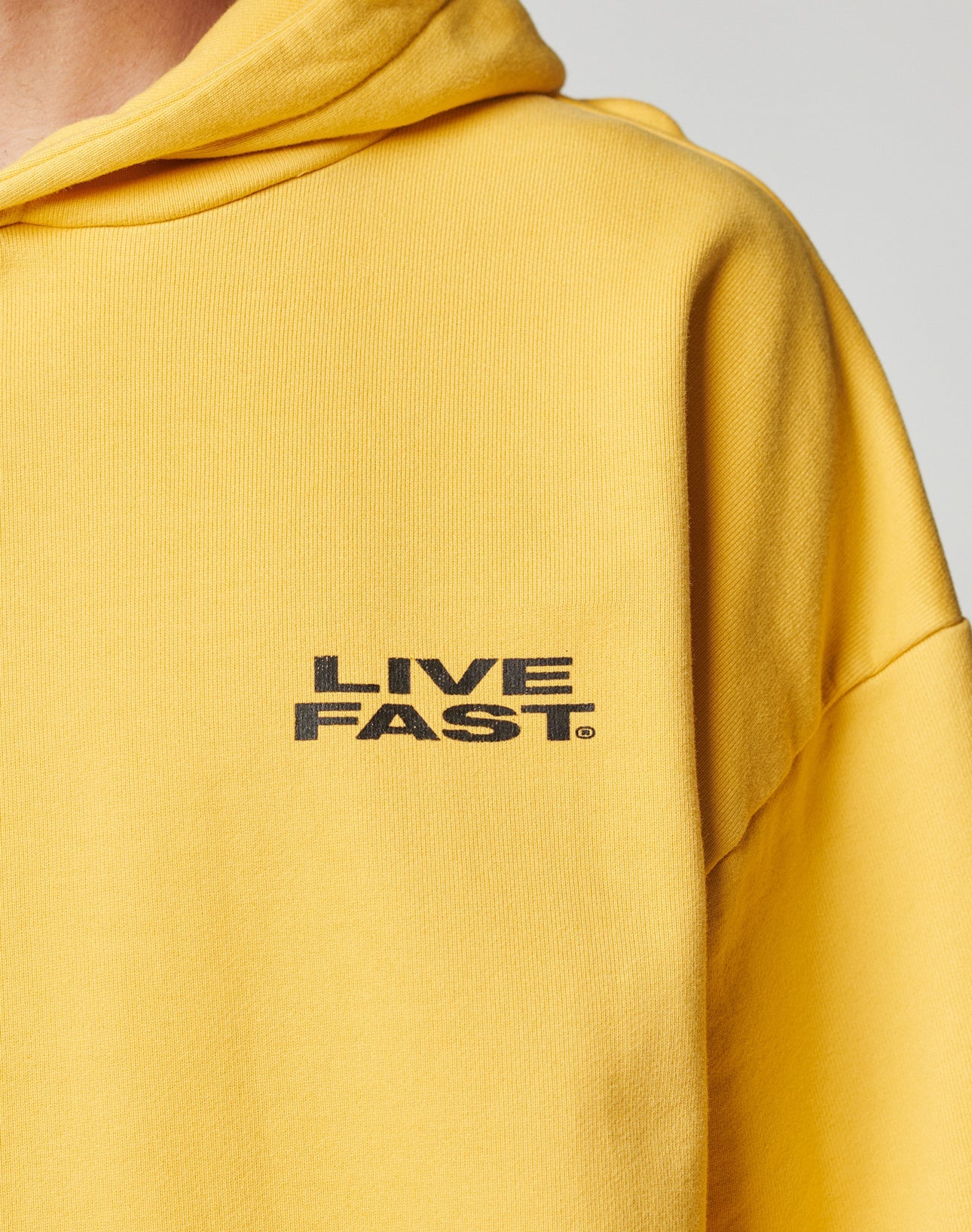 Close-up of a person wearing the yellow Print Over Hooded sweatshirt by LIVE FAST DIE YOUNG with LIVE FAST. printed in black on the chest. Only the upper body and part of the hoodie are seen against a neutral background.