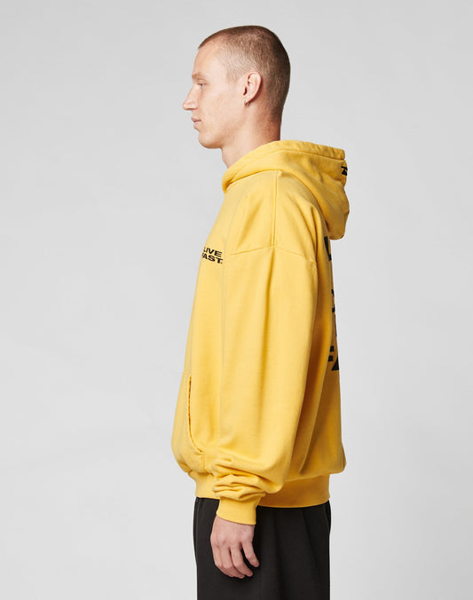 A person with short hair stands in profile, wearing a yellow LIVE FAST DIE YOUNG Print Over Hooded sweatshirt made from 100% Baumwolle and black pants against a plain light gray background.