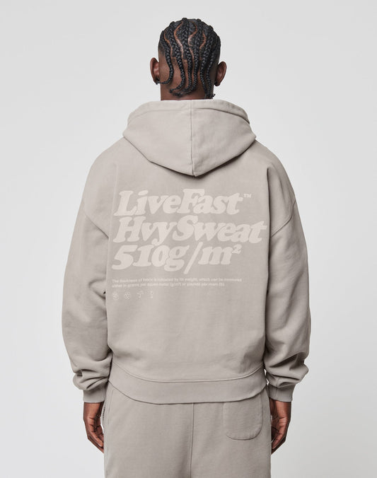 A person with short, braided hair is seen from behind, wearing the LIVE FAST DIE YOUNG Basic 510 Hooded Zip in light taupe, featuring white LiveFast™ HvySweat 510g/m² and small text printed on the back.