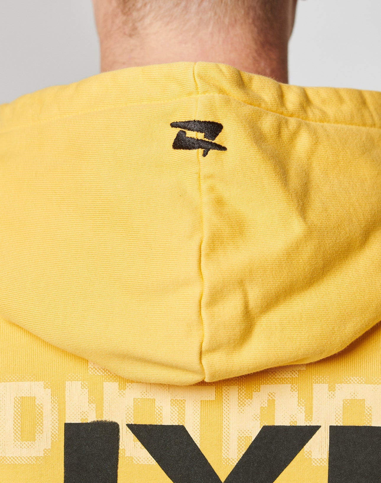 Close-up of the back of a person wearing the LIVE FAST DIE YOUNG Print Over Hooded, an oversized yellow hoodie with a small black logo at the top center and large black-and-white print on the front and back.