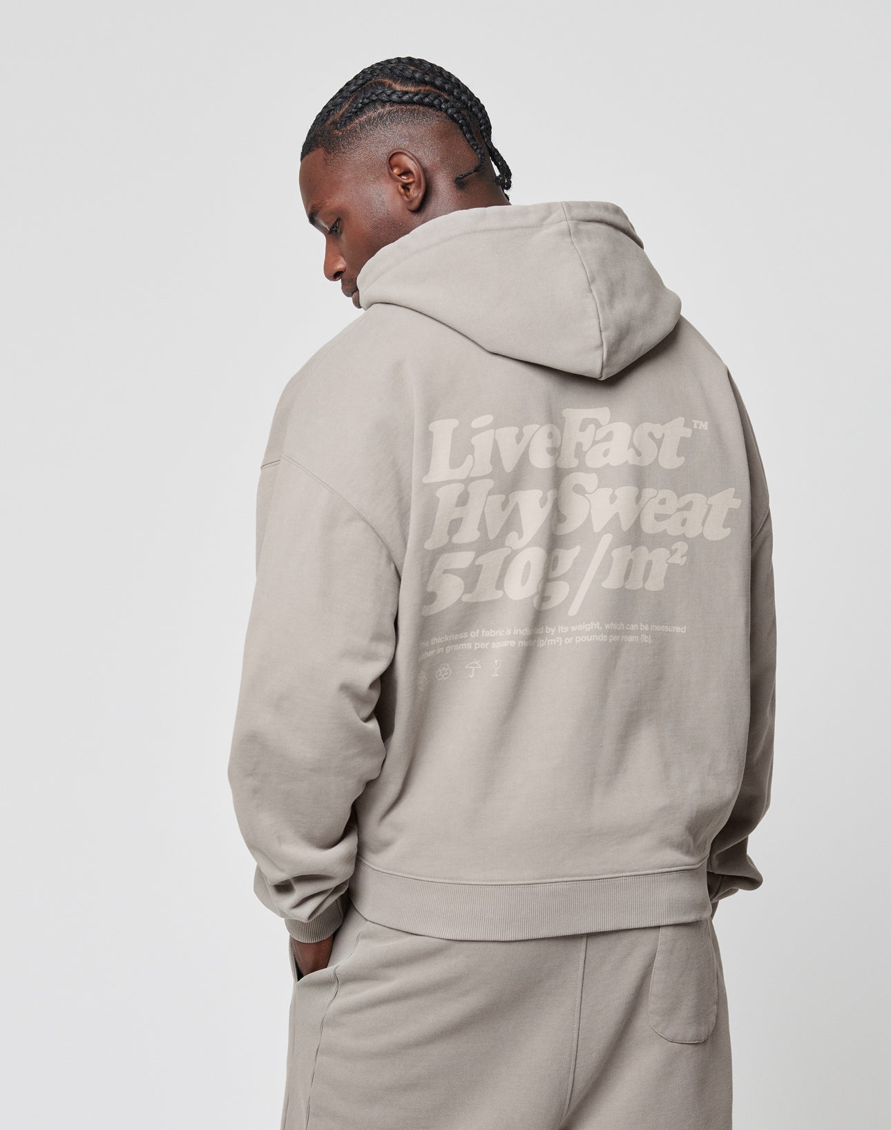 A person with braided hair, seen from the side and back, wears a beige LIVE FAST DIE YOUNG Basic 510 Hooded Zip in an oversize fit. LiveFast Hvy Sweat 510g/m² is printed large on the back against a plain white background.