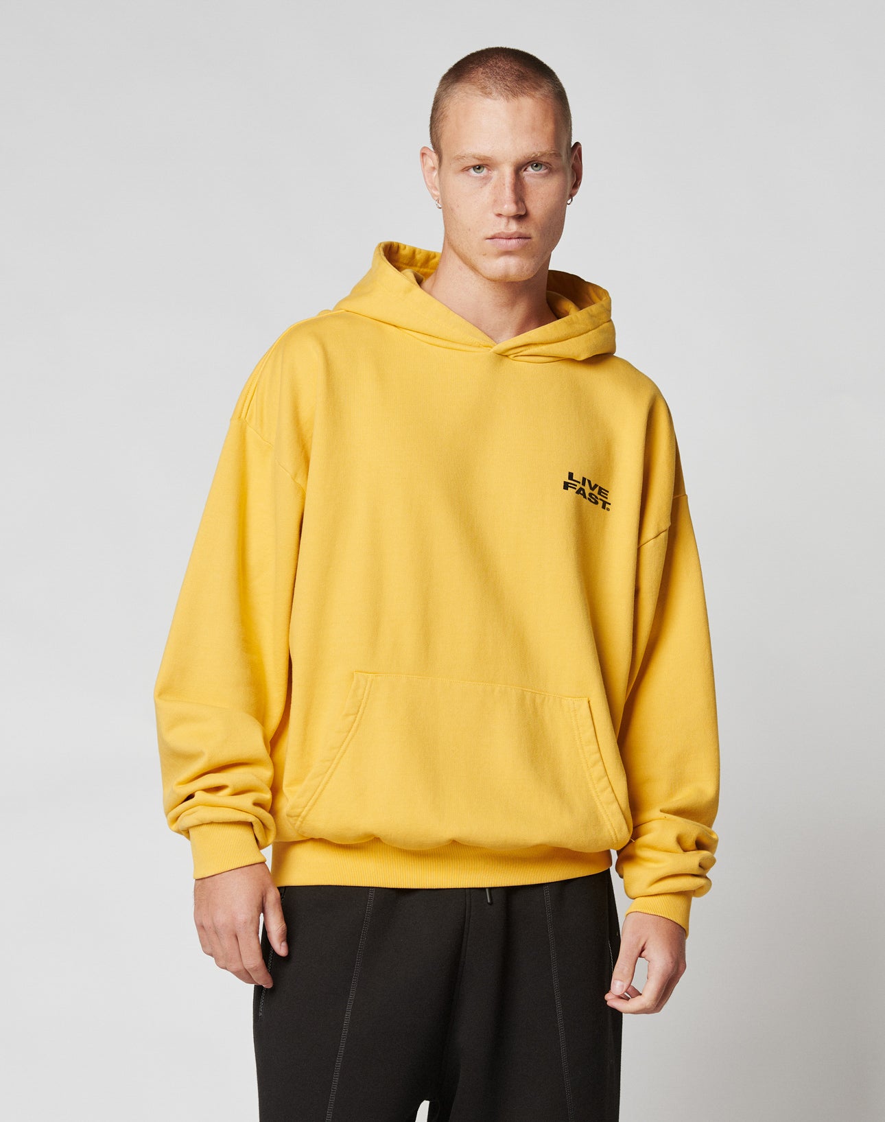 Wearing the Print Over Hooded by LIVE FAST DIE YOUNG, a person in an oversize yellow hoodie with black chest text and a front pocket stands against a light gray background. The hoodie is made from 100% Baumwolle for comfort.