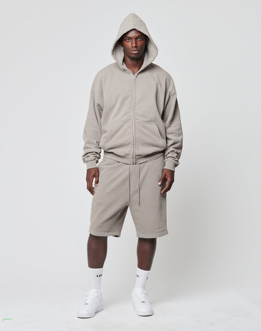A man stands facing forward wearing LIVE FAST DIE YOUNG Basic Heavy Shorts, a taupe logo-print hooded zip-up sweatshirt, white crew socks, and white sneakers against a plain light background.