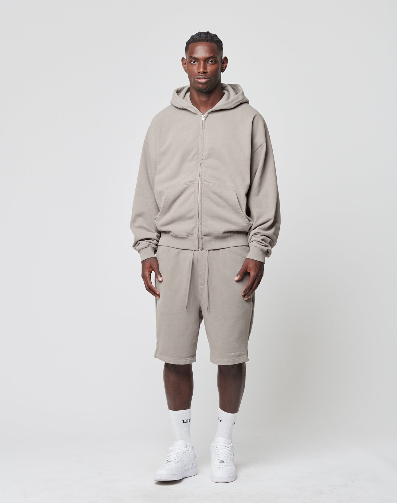 A man faces forward with a neutral expression, wearing the LIVE FAST DIE YOUNG Basic 510 Hooded Zip in light gray Oversize Fit, matching shorts, white crew socks, and white sneakers against a plain white background.