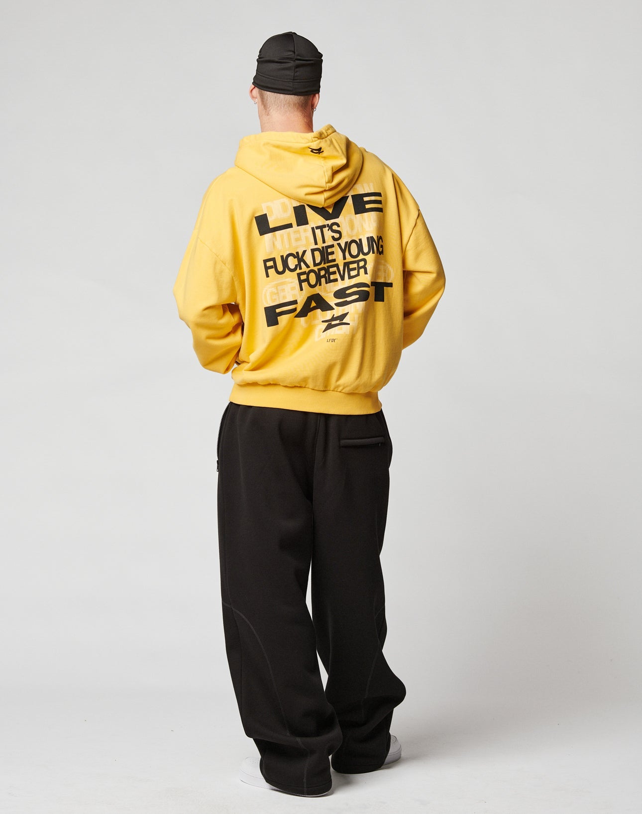 A person models the LIVE FAST DIE YOUNG Print Over Hooded in yellow, featuring bold black front and back prints, styled with black wide-leg pants, white shoes, and a black cap against a plain light backdrop.