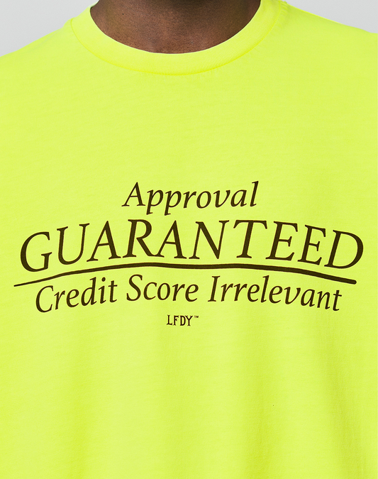 A person wears the LFDY Real Est.8 Longsleeve by LIVE FAST DIE YOUNG, an oversized bright yellow shirt with "Approval GUARANTEED Credit Score Irrelevant LFDY" boldly printed in black on the front, made from 100% Baumwolle for comfort.