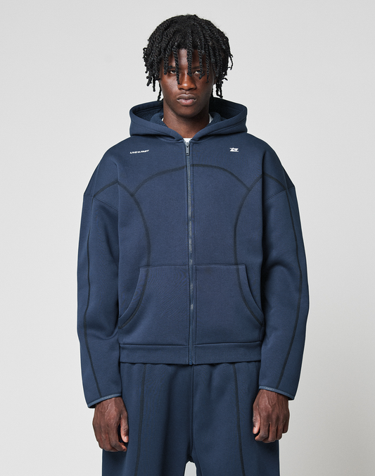 A person with medium-length, textured hair wears the LIVE FAST DIE YOUNG LFDY Active Fleece Hooded Zip and matching pants in navy blue, standing against a plain light gray background and looking straight at the camera. | model