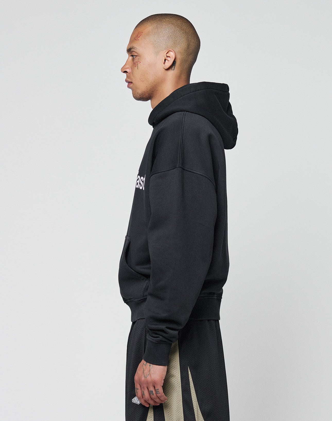 A person with a shaved head and facial tattoos stands in profile against a plain background, wearing the LIVE FAST DIE YOUNG Basic +49 Hooded sweatshirt and black shorts with beige accents.