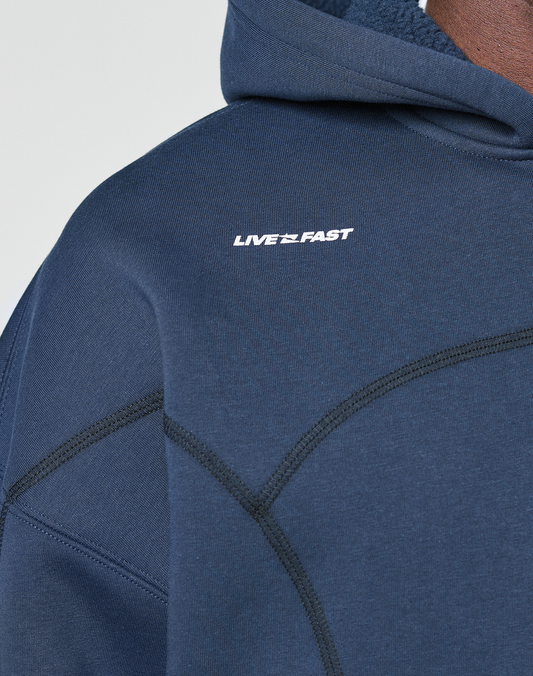 Close-up of the LFDY Active Fleece Hooded Zip by LIVE FAST DIE YOUNG, featuring an oversize navy blue fit in cotton with black stitching and a white LIVE FAST logo on the chest. Part of the wearer's neck and chin are visible.