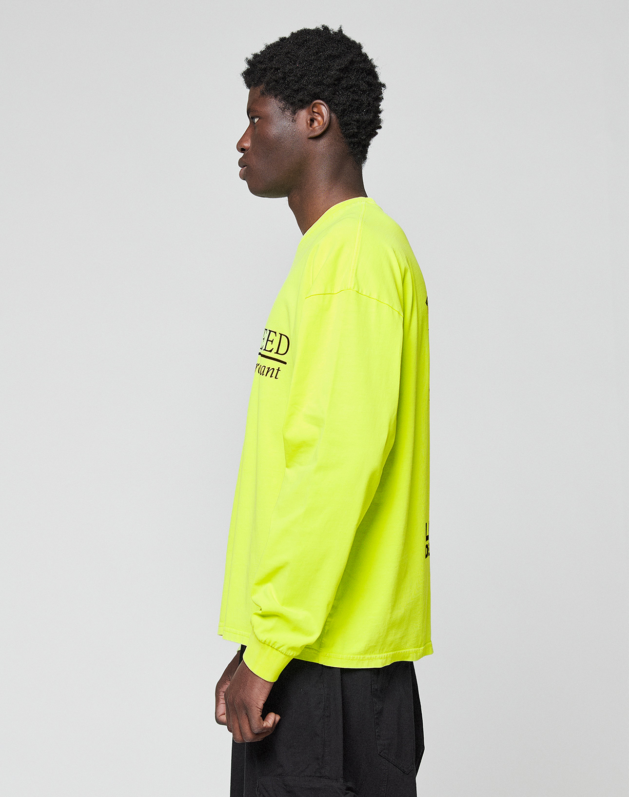 A person stands in profile against a plain background, wearing the LIVE FAST DIE YOUNG LFDY Real Est.8 Longsleeve in neon yellow with black front and back print, paired with black pants. Made of 100% Baumwolle.