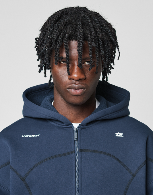 A person with short, twisted hair wears the LIVE FAST DIE YOUNG LFDY Active Fleece Hooded Zip in navy blue, featuring a subtle logo print, looking neutrally at the camera against a plain light background.