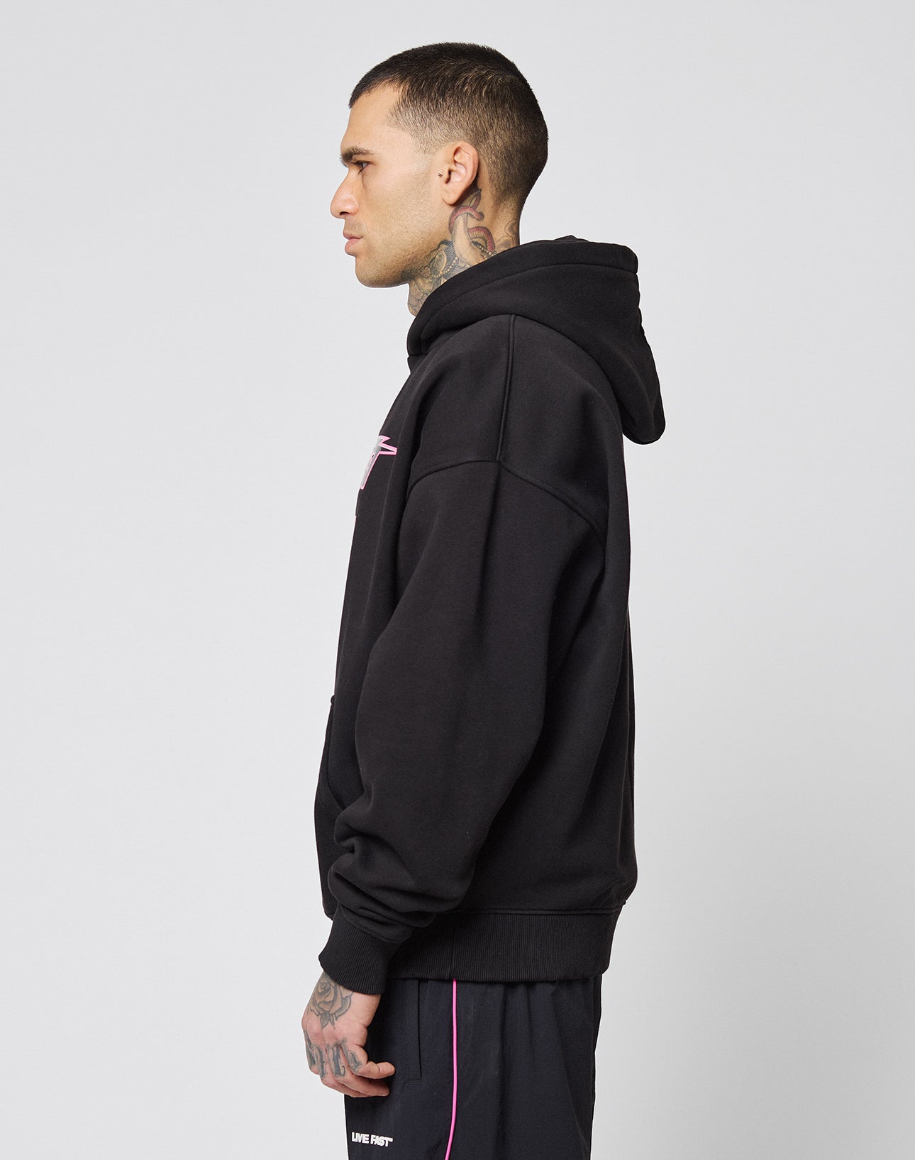 A tattooed man in profile wears a black LIVE FAST DIE YOUNG LFDY Triple Racing Hooded in Oversize Fit and black shorts with pink trim, standing against a plain light gray background.