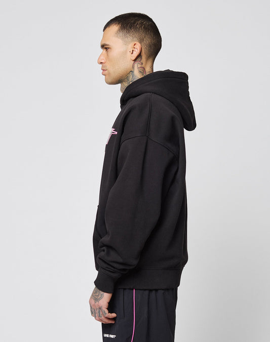 A tattooed man in profile wears a black LIVE FAST DIE YOUNG LFDY Triple Racing Hooded in Oversize Fit and black shorts with pink trim, standing against a plain light gray background.