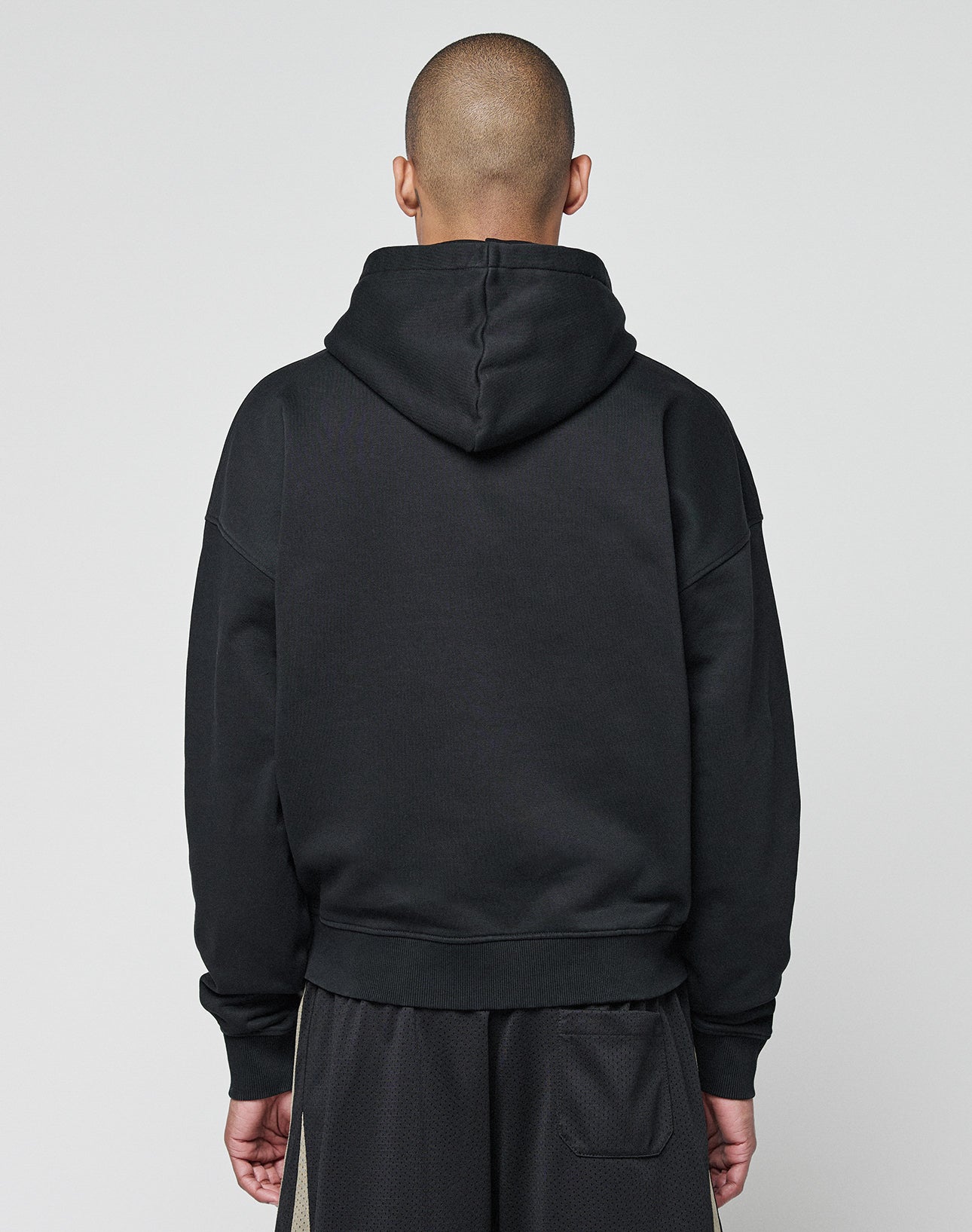 A person with a shaved head faces away from the camera, wearing the LIVE FAST DIE YOUNG Basic +49 Hooded in oversize fit and matching black Baumwolle Polyester shorts, set against a plain light gray background.