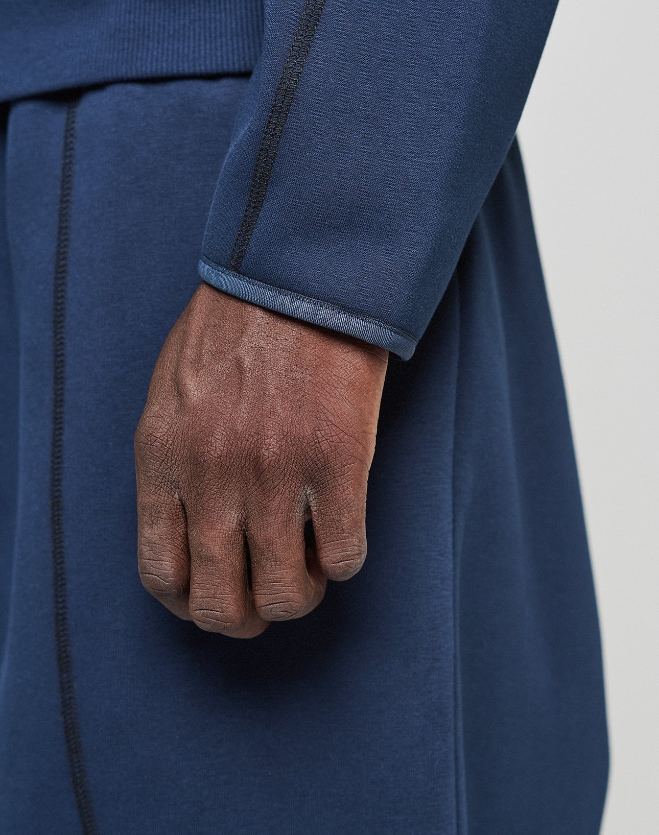A close-up of a hand and forearm wearing the LIVE FAST DIE YOUNG LFDY Active Fleece Hooded Zip in navy blue, featuring an oversize fit and black stitching. Matching pants are shown against a neutral, blurred background.