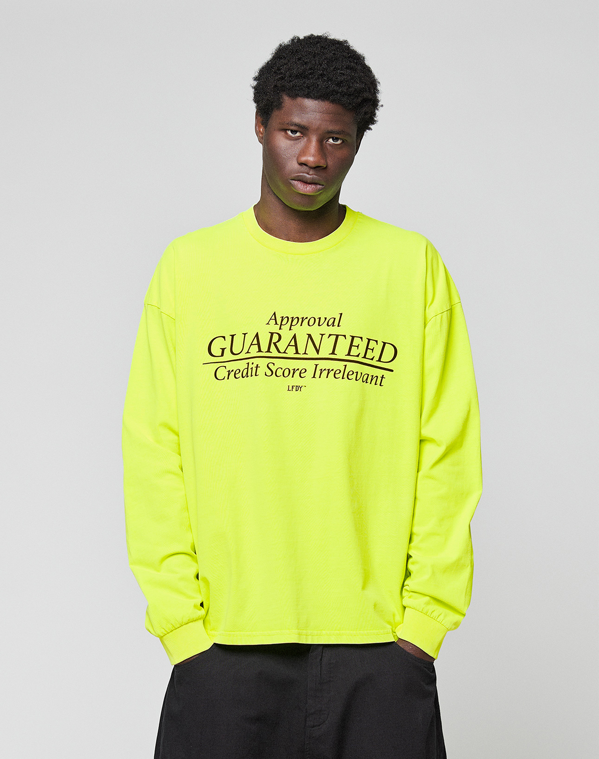 Wearing the LFDY Real Est.8 Longsleeve by LIVE FAST DIE YOUNG, an oversize neon yellow 100% Baumwolle shirt with black "Approval GUARANTEED Credit Score Irrelevant" prints, a person stands against a plain light gray background.