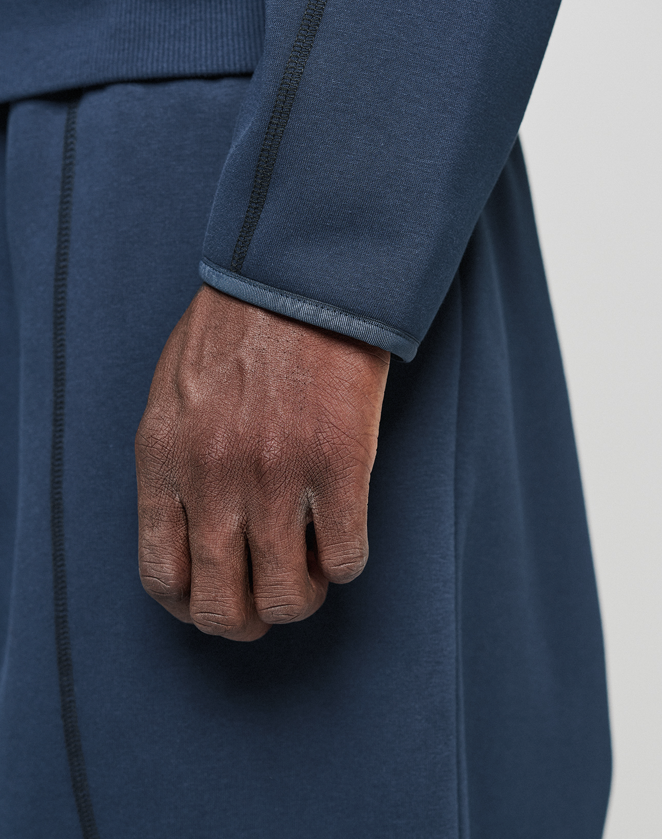 A close-up of a medium-brown hand by their side, wearing the LIVE FAST DIE YOUNG LFDY Active Fleece Hooded Zip in dark blue with black seam stitching and matching pants.