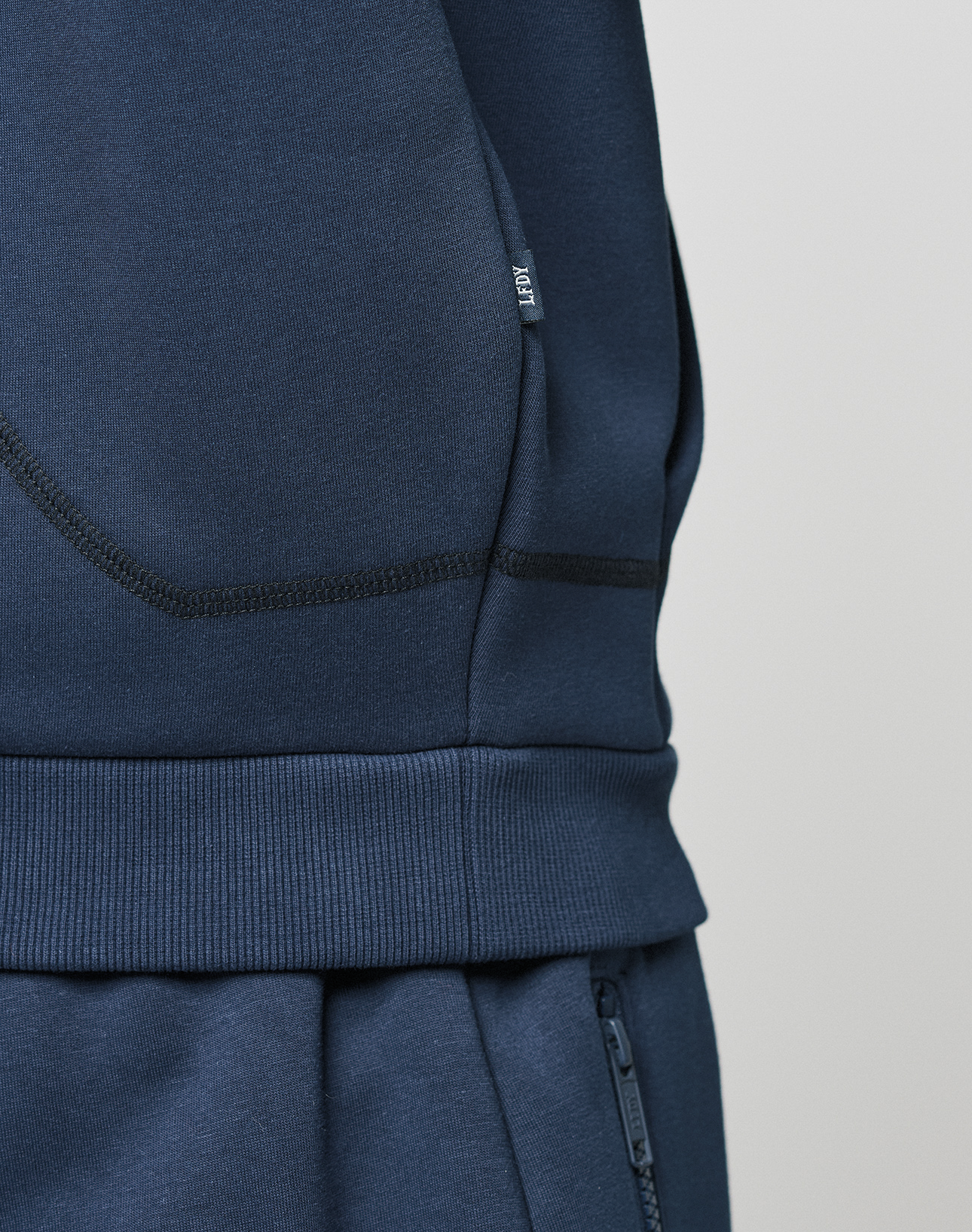 Close-up of a person in LIVE FAST DIE YOUNG's LFDY Active Fleece Hooded Zip, navy blue, oversize fit. The image highlights the hoodie’s pocket and waistband details, with visible stitching, a fabric tag, and subtle logo print.