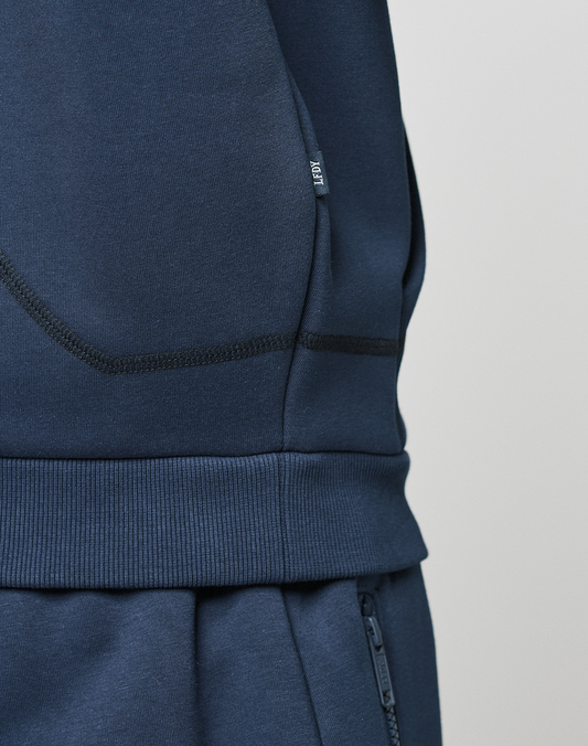 Close-up of a person in LIVE FAST DIE YOUNG's LFDY Active Fleece Hooded Zip, navy blue, oversize fit. The image highlights the hoodie’s pocket and waistband details, with visible stitching, a fabric tag, and subtle logo print.