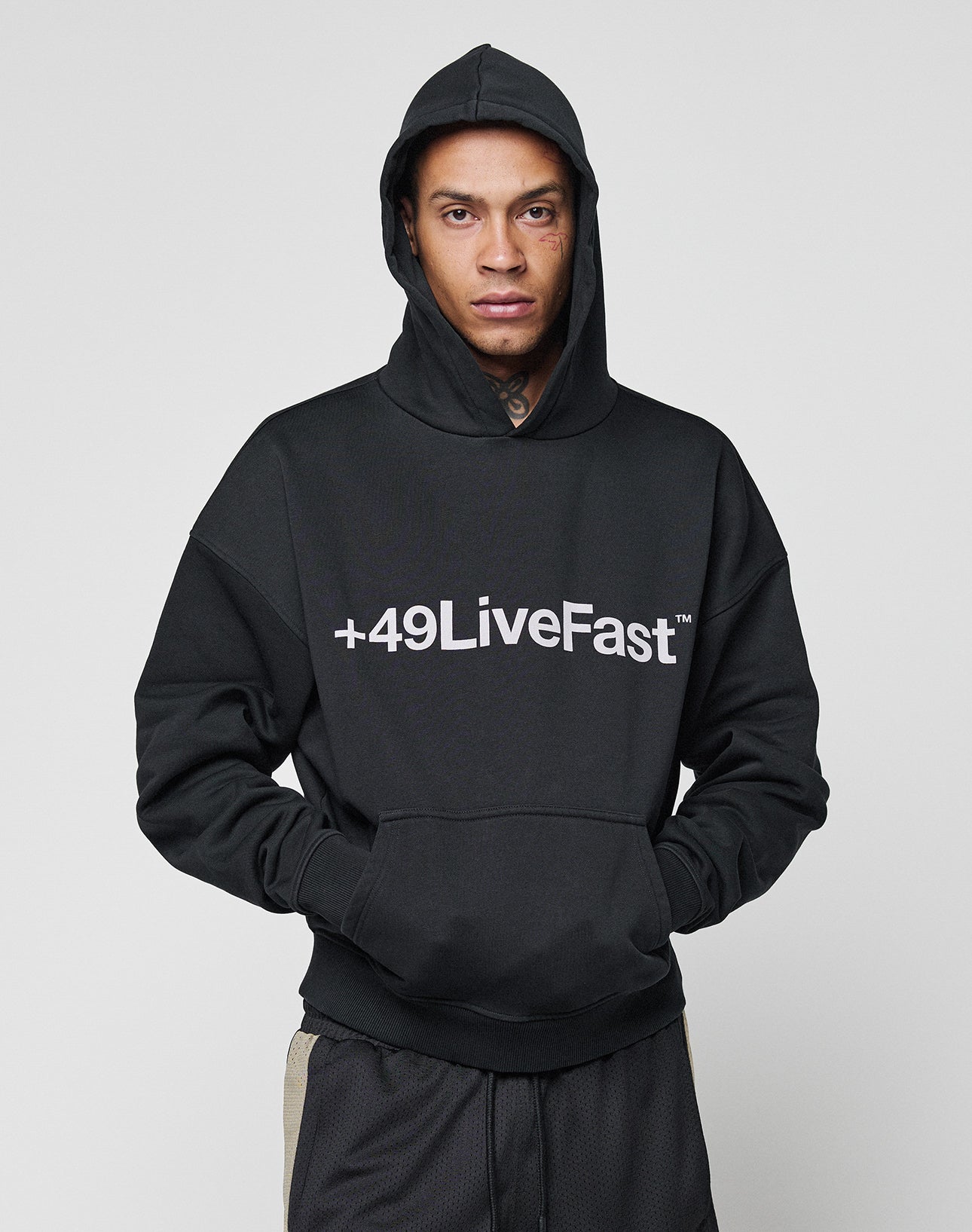 A person wears the Basic +49 Hooded by LIVE FAST DIE YOUNG, an oversized black hoodie with +49LiveFast front print, hands in pockets and hood up, standing against a plain light background. Crafted from a comfy cotton-polyester blend.