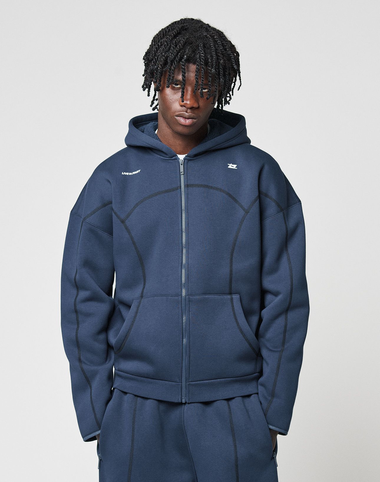 A young man with short, textured dreadlocks wears the navy LFDY Active Fleece Hooded Zip and matching pants by LIVE FAST DIE YOUNG, featuring a subtle logo print and crafted from soft Baumwolle, standing against a plain light gray background.