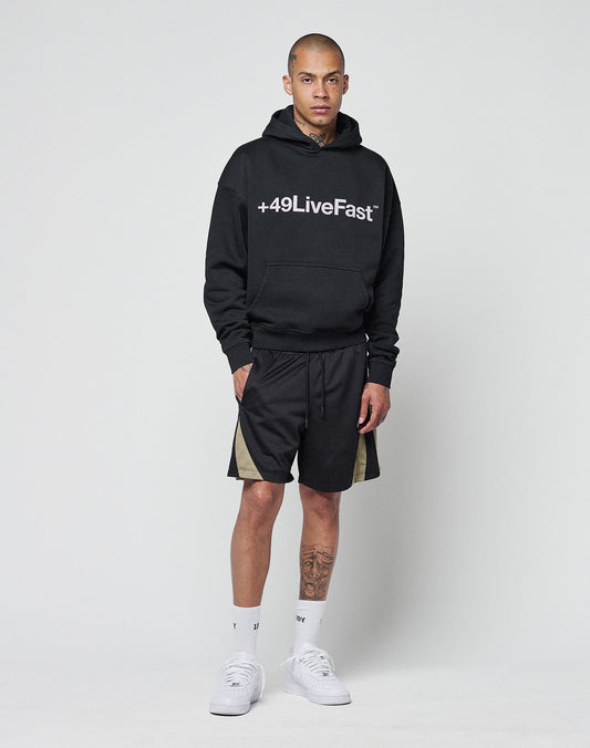 A man wears the LIVE FAST DIE YOUNG Basic +49 Hooded in black, featuring a +49LiveFast front print and oversize fit (Baumwolle-Polyester blend), paired with black shorts with beige side panels, white socks, and white sneakers.