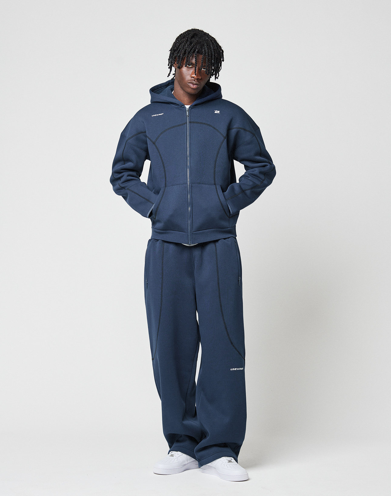 A person with short, twisted hair wears the LIVE FAST DIE YOUNG LFDY Active Fleece Hooded Zip in dark blue and matching wide-leg pants with white sneakers, standing against a plain white background with hands in their hoodie pockets.