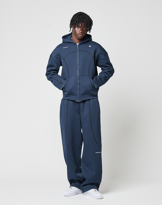 A person with short, twisted hair wears the LIVE FAST DIE YOUNG LFDY Active Fleece Hooded Zip in dark blue and matching wide-leg pants with white sneakers, standing against a plain white background with hands in their hoodie pockets.