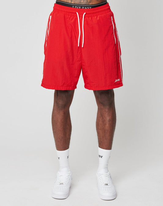 A person wears bright red LIVE FAST DIE YOUNG Beach Shorts with an elastic waistband and white drawstrings, paired with white socks and sneakers, standing against a plain light gray background. Only the lower half is visible.