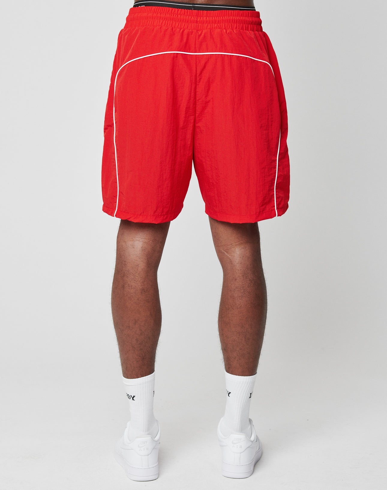 A person faces away, wearing LIVE FAST DIE YOUNG Beach Shorts in bright red with white piping and an elastic waistband, paired with white crew socks and sneakers, all set against a plain white background.