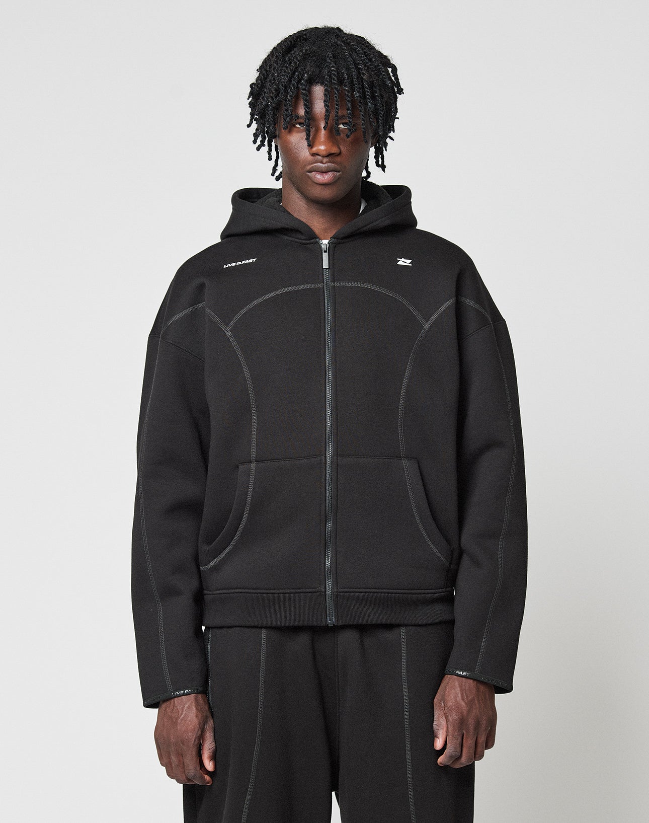 A person with medium-length twisted hair wears the LIVE FAST DIE YOUNG LFDY Active Fleece Hooded Zip in black, styled oversized with matching black pants, standing against a plain light gray background and looking at the camera. | model