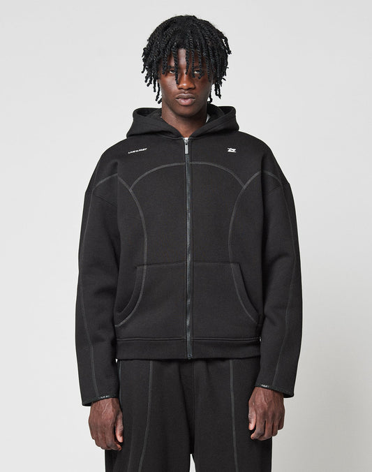 A person with medium-length twisted hair wears the LIVE FAST DIE YOUNG LFDY Active Fleece Hooded Zip in black, styled oversized with matching black pants, standing against a plain light gray background and looking at the camera. | model