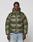 A man wears the LIVE FAST DIE YOUNG Live Fast Puffer Jacket in olive green with nylon outer material, oversized fit, hood, and a reflective LIVE FAST logo, styled with light blue jeans against a plain background.
