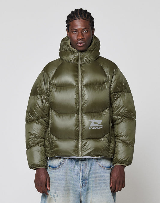 A man wears the LIVE FAST DIE YOUNG Live Fast Puffer Jacket in olive green with nylon outer material, oversized fit, hood, and a reflective LIVE FAST logo, styled with light blue jeans against a plain background.