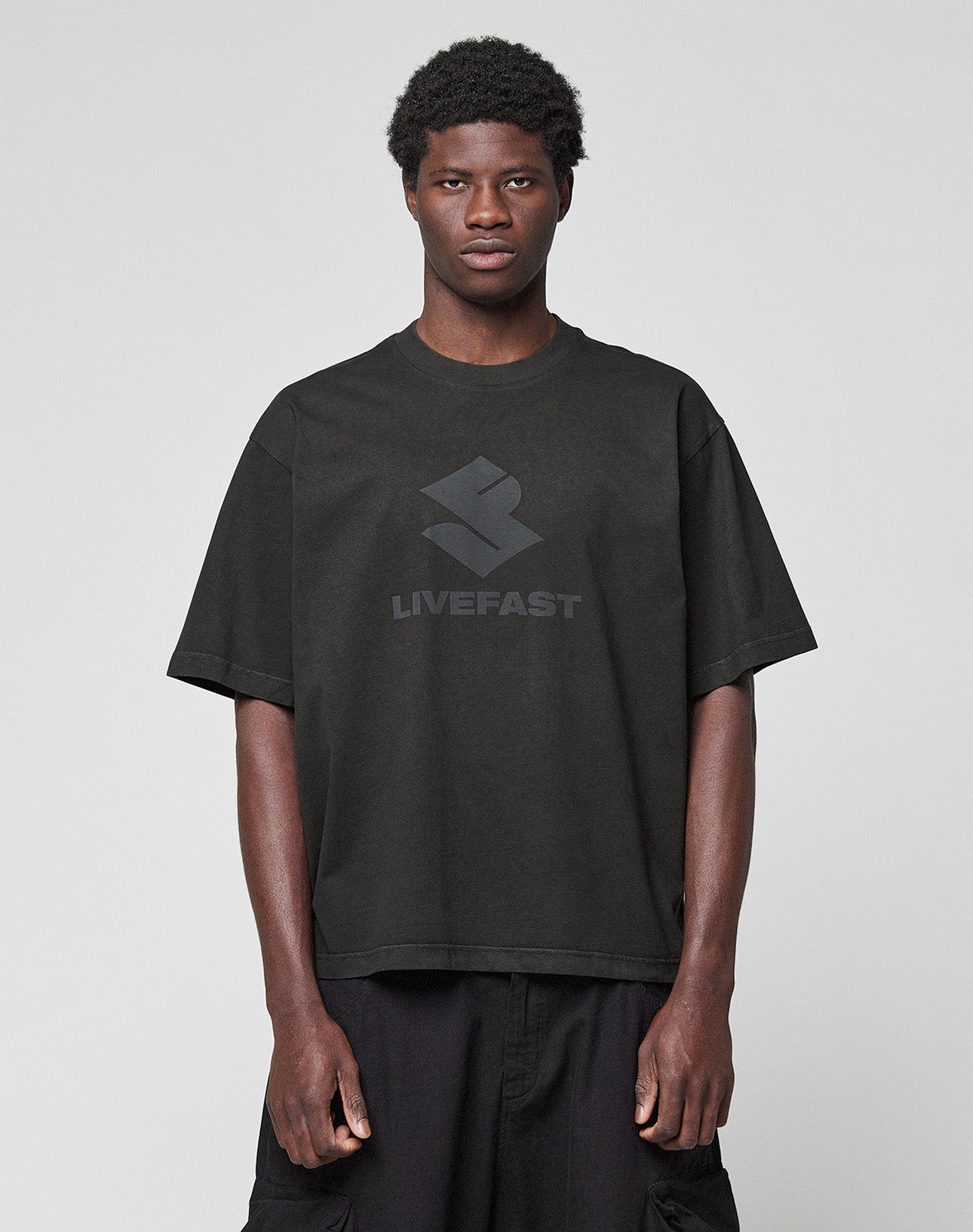 A person stands against a light gray background, wearing the Live Fast LF Tee by LIVE FAST DIE YOUNG—an oversize-fit black T-shirt with a "LIVEFAST" front print, made from 100% Baumwolle—and paired with black shorts.