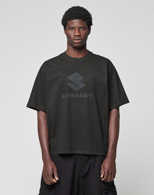 A person stands against a light gray background, wearing the Live Fast LF Tee by LIVE FAST DIE YOUNG—an oversize-fit black T-shirt with a "LIVEFAST" front print, made from 100% Baumwolle—and paired with black shorts. | model