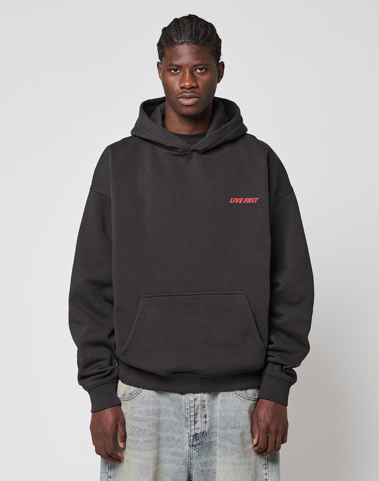 A person faces forward in a LIVE FAST DIE YOUNG LFDY Globe Hooded black hoodie with red LIVE FAST text on the left chest, made of cotton-polyester, and light blue jeans. The background is plain light gray.