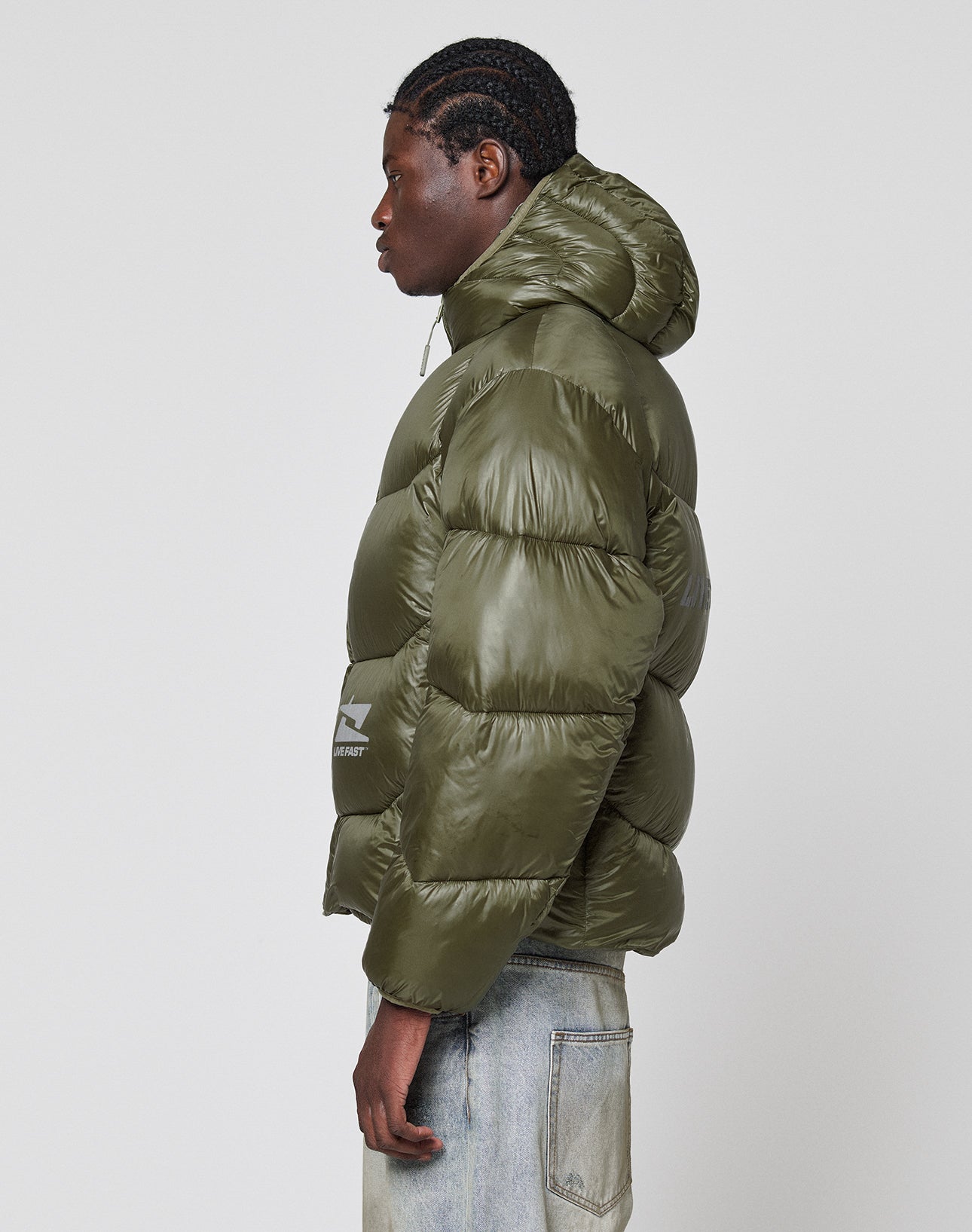 A person with braided hair stands in profile, wearing an olive green Live Fast Puffer Jacket by LIVE FAST DIE YOUNG, paired with light blue jeans against a plain white background.