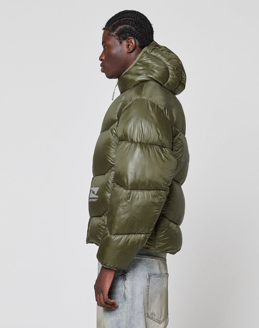 A person with braided hair stands in profile, wearing an olive green Live Fast Puffer Jacket by LIVE FAST DIE YOUNG, paired with light blue jeans against a plain white background.