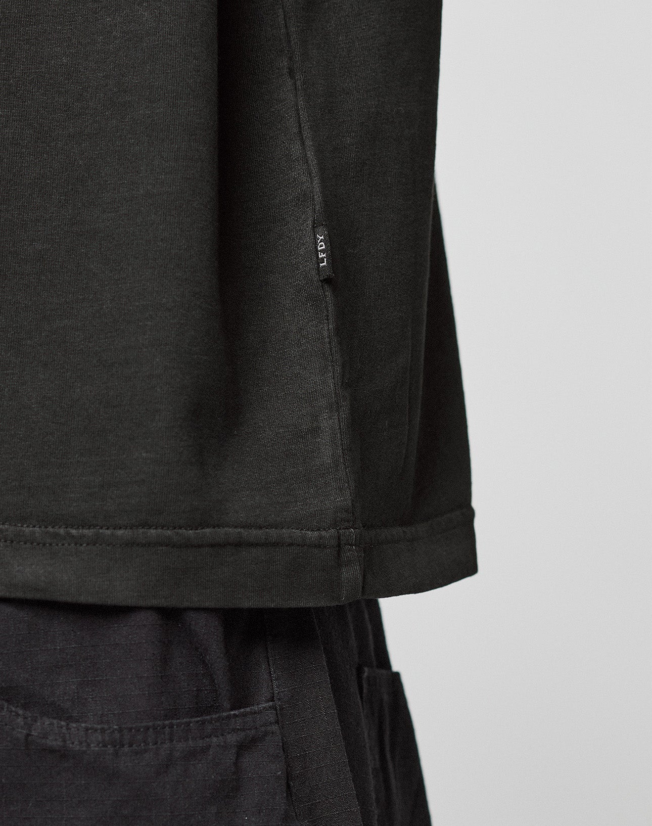 Close-up of a person wearing the Live Fast LF Tee by LIVE FAST DIE YOUNG, an oversized black shirt with black pants. A small fabric tag is attached near the hem. The background is plain and light-colored.
