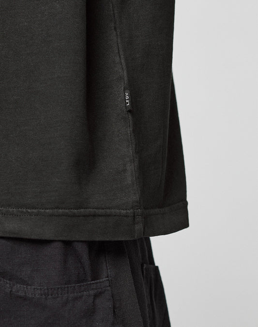 Close-up of a person wearing the Live Fast LF Tee by LIVE FAST DIE YOUNG, an oversized black shirt with black pants. A small fabric tag is attached near the hem. The background is plain and light-colored.
