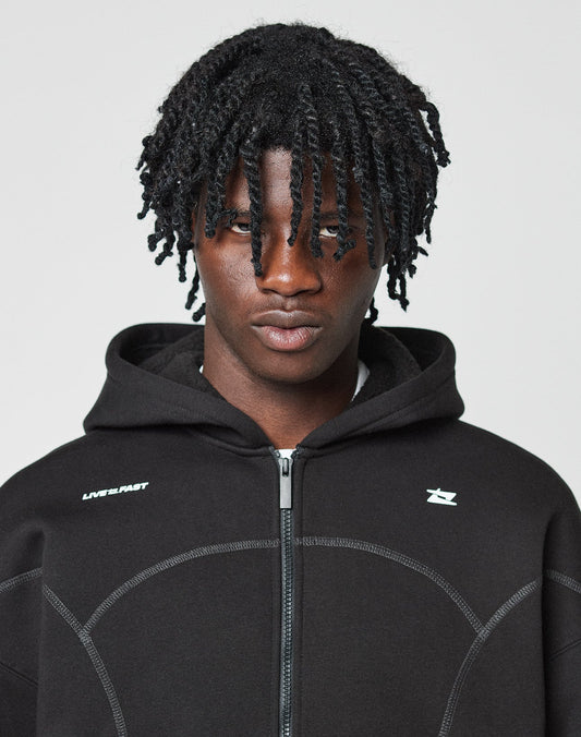 A person with medium-dark skin and tightly coiled hair looks at the camera, wearing the LIVE FAST DIE YOUNG LFDY Active Fleece Hooded Zip—an oversize black hoodie with a white logo on the chest—against a plain light background.