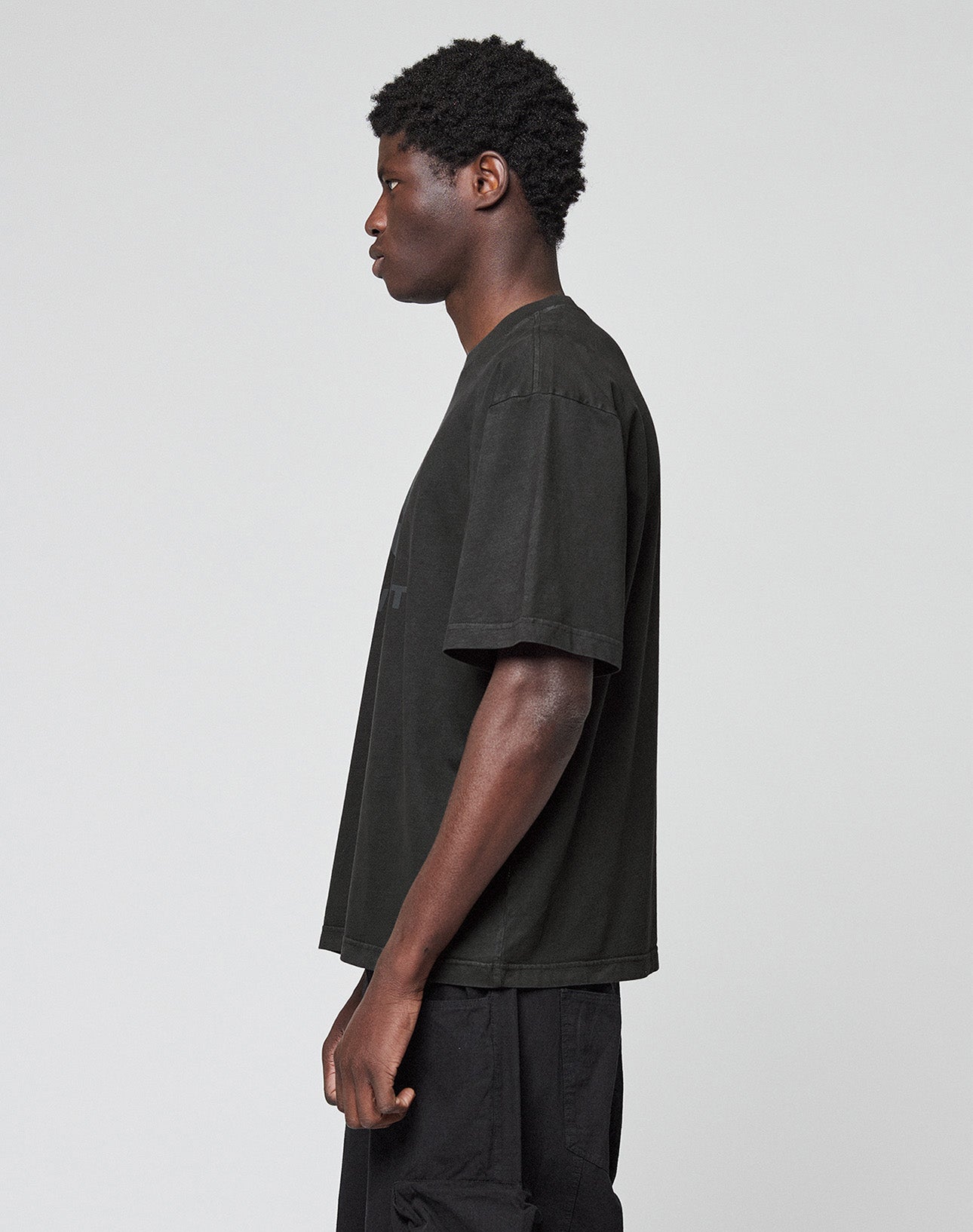 A person with short curly hair stands sideways, facing left, wearing the LIVE FAST DIE YOUNG Live Fast LF Tee in Oversize Fit and black pants against a plain light gray background.