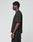 A person with short curly hair stands sideways, facing left, wearing the LIVE FAST DIE YOUNG Live Fast LF Tee in Oversize Fit and black pants against a plain light gray background.