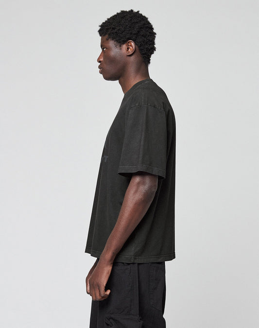 A person with short curly hair stands sideways, facing left, wearing the LIVE FAST DIE YOUNG Live Fast LF Tee in Oversize Fit and black pants against a plain light gray background.