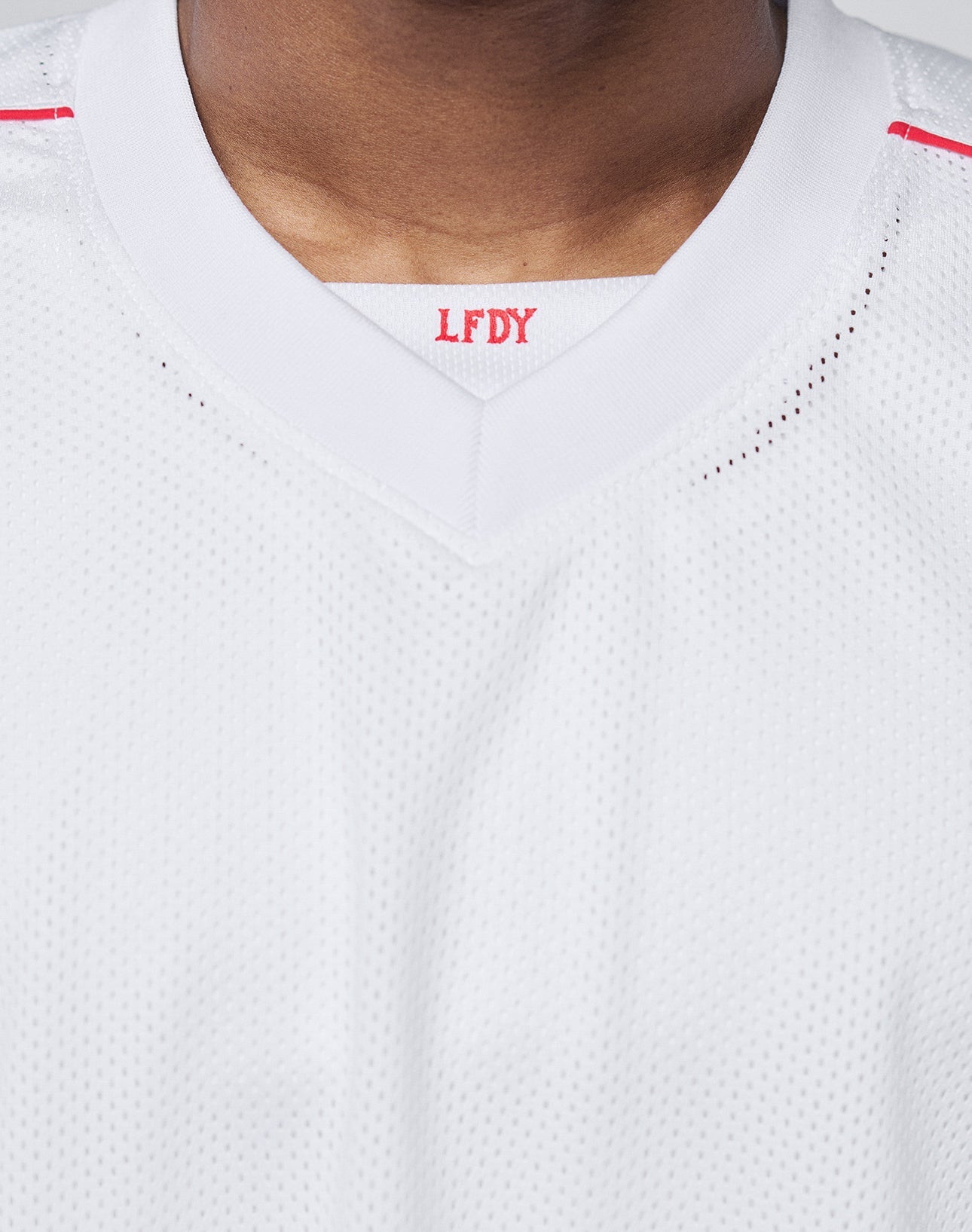 Close-up of a person wearing the Basic Mesh Jersey by LIVE FAST DIE YOUNG. This white oversize-fit sports jersey is made from 100% polyester with a V-neck collar. Red LFDY letters are visible inside the collar; face not shown.