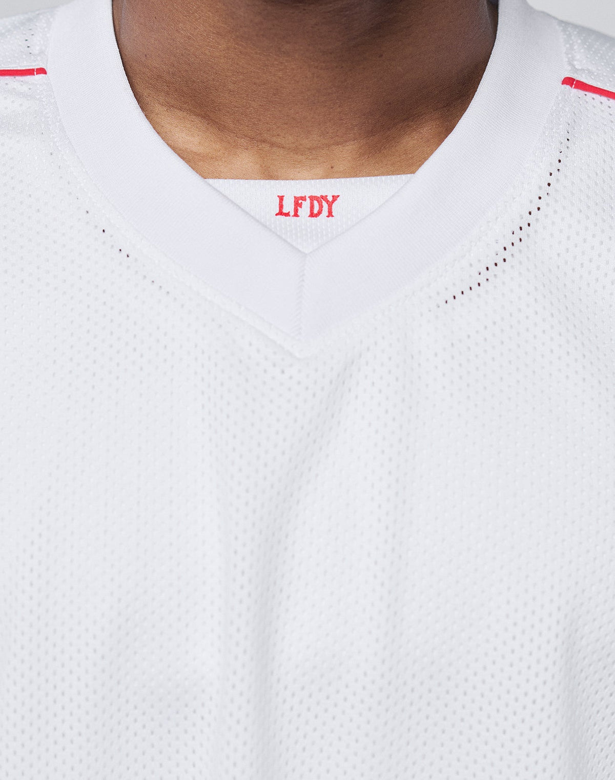 Close-up of a person wearing the Basic Mesh Jersey by LIVE FAST DIE YOUNG. This white oversize-fit sports jersey is made from 100% polyester with a V-neck collar. Red LFDY letters are visible inside the collar; face not shown.
