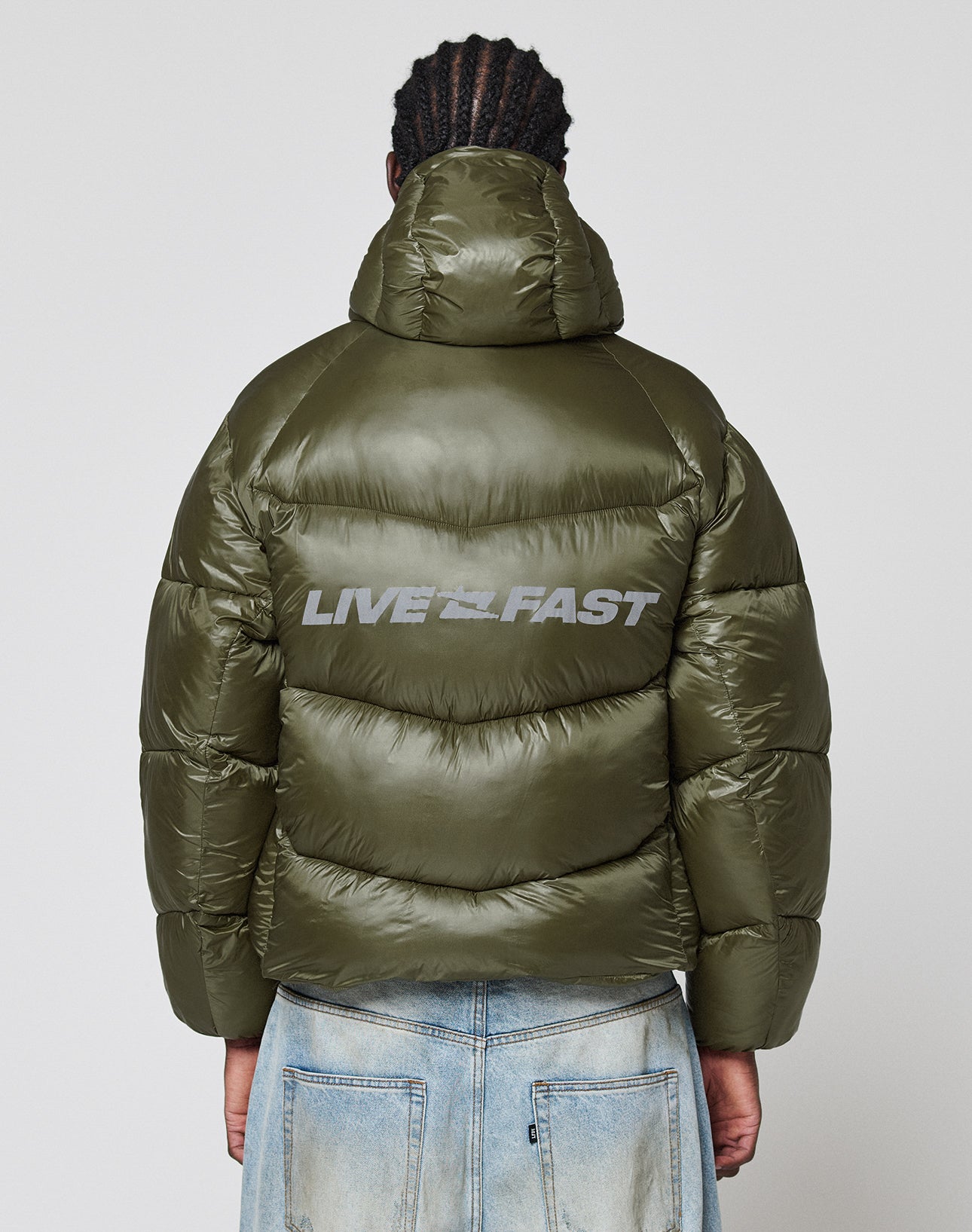 A person stands with their back to the camera, wearing the LIVE FAST DIE YOUNG Live Fast Puffer Jacket—an oversize fit with shiny olive green nylon and a bold, reflective "LIVE FAST" print on the back—against a plain white background.