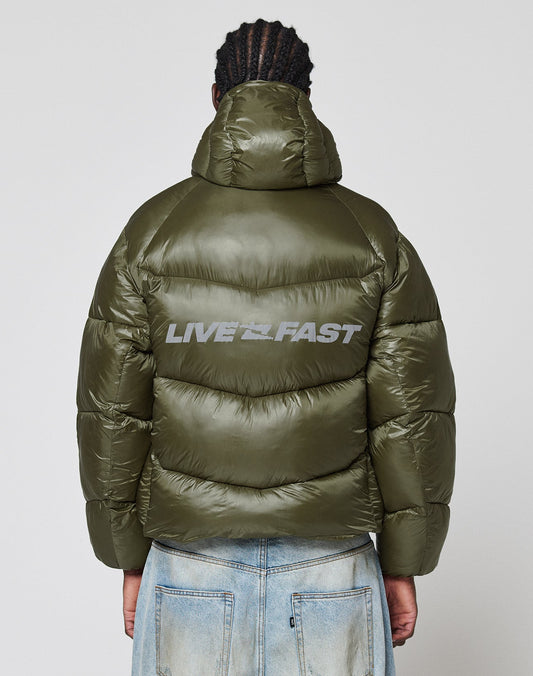 A person stands with their back to the camera, wearing the LIVE FAST DIE YOUNG Live Fast Puffer Jacket—an oversize fit with shiny olive green nylon and a bold, reflective "LIVE FAST" print on the back—against a plain white background.