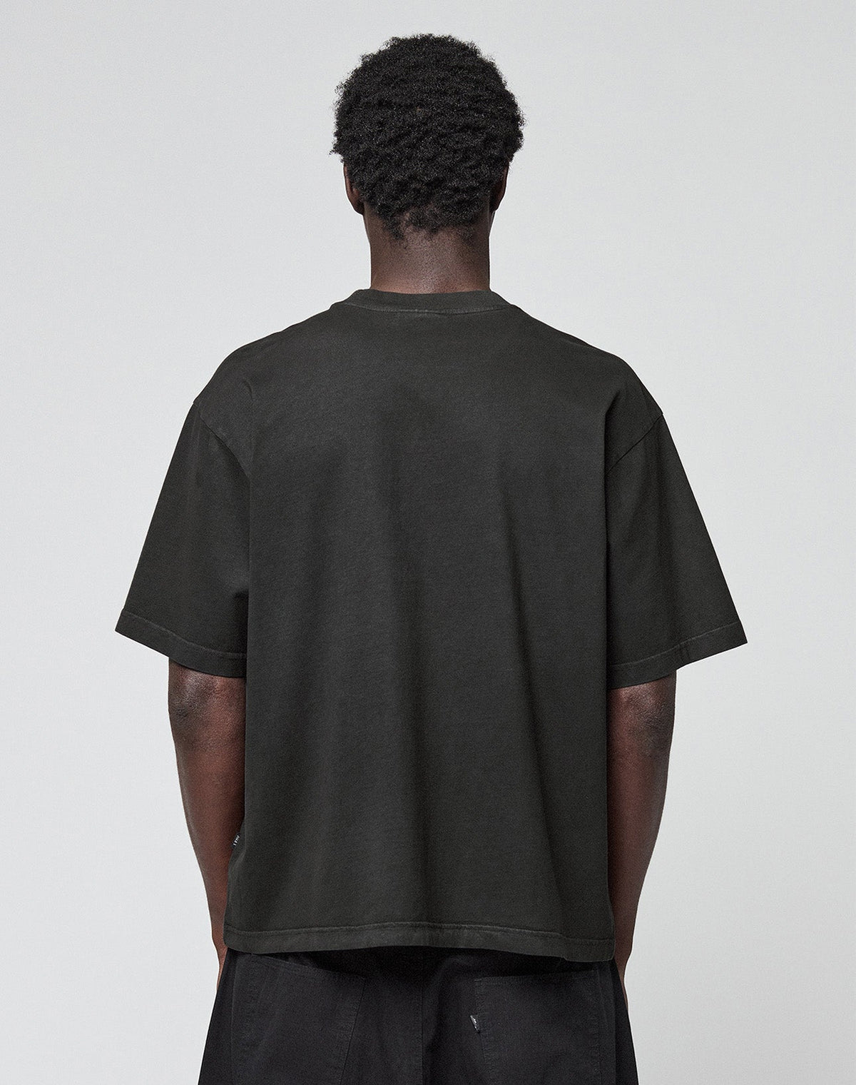 A person with short curly hair stands with their back to the camera, wearing the LIVE FAST DIE YOUNG Live Fast LF Tee in black and matching black pants against a plain light gray background.